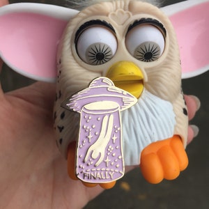 Op de afbeelding: Een gouden emaille pin met een paarse UFO en een hand die eruit reikt. De pin heeft de tekst "FINALLY" onderaan. De pin is bevestigd aan een wit en oranje pluche speelgoed.
