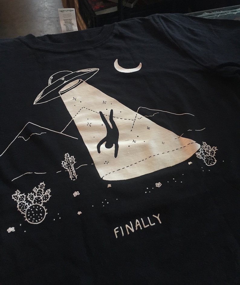 May include: Black t-shirt with a white graphic of an alien spaceship beaming a person up from a desert landscape. The text "FINALLY" is printed below the graphic.