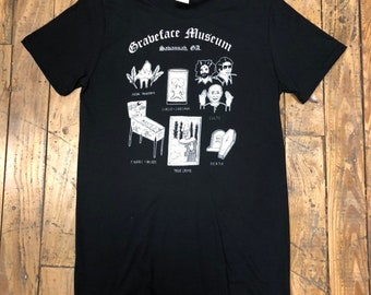 May include: Black t-shirt with a white graphic of the Graveface Museum in Savannah, GA. The graphic features illustrations of a pinball machine, a true crime scene, a cult, a pair of conjoined twins, and a coffin.
