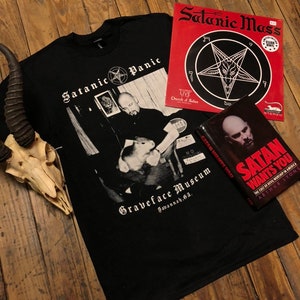 May include: A black t-shirt with a white graphic print of a man holding a small animal. The graphic includes the text "Satanic Panic" and "Graveface Museum Savannah, GA".