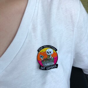 Pode incluir: Um broche de esmalte preto com um esqueleto de desenho animado comendo lixo. O broche tem um fundo rosa, laranja e amarelo com o texto "Stop Pollution Eat Garbage".