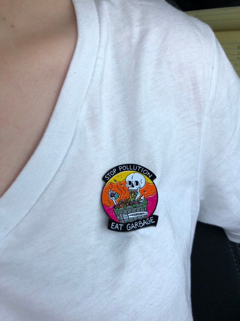 Pode incluir: Um broche de esmalte preto com um esqueleto de desenho animado comendo lixo. O broche tem um fundo rosa, laranja e amarelo com o texto "Stop Pollution Eat Garbage".