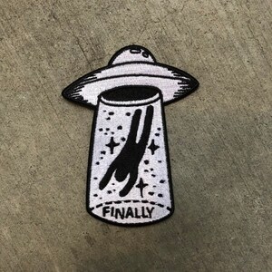 Op de afbeelding: Geborduurde patch met een zwart-wit UFO-ontwerp. Een stokfiguur wordt ontvoerd door de UFO, met het woord "FINALLY" onderaan. Geschikt voor kleding of accessoires.