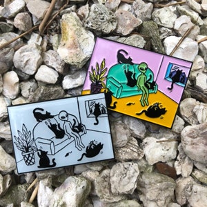 May include: Two enamel pins featuring a cartoon alien and black cats in a living room setting. One pin has a pink background with a green couch, the other has a light blue background. The pins are set against a background of rocks.