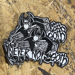 May include: Black and white embroidered patches with the text "NEVER NOT GOTH" and a stylized illustration of a person lying in a coffin. The patches are stacked on a wood-grain surface.