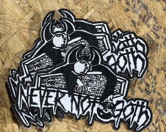 May include: Black and white embroidered patches with the text "NEVER NOT GOTH" and a stylized illustration of a person lying in a coffin. The patches are stacked on a wood-grain surface.