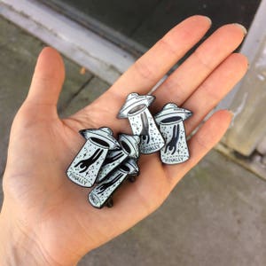 Op de afbeelding: Een verzameling zwart-witte emaille pins in de vorm van vliegende schotels. Elke pin heeft een gestileerde alien figuur en het woord "FINALLY". De pins worden in een hand gehouden.