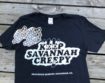 May include: Black t-shirt with a white graphic that says "Keep Savannah Creepy" with a claw hand design. The shirt also has a smaller sticker that says "Keep Savannah Creepy" on the front. The shirt is on a wooden surface.