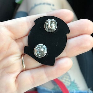 Pode incluir: O verso de um broche de esmalte preto com dois fechos de metal. O broche tem a forma de uma letra Q estilizada.