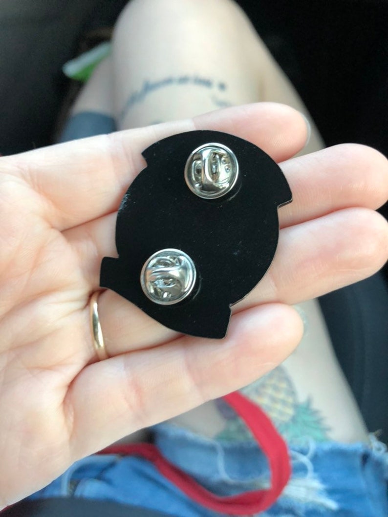 Pode incluir: O verso de um broche de esmalte preto com dois fechos de metal. O broche tem a forma de uma letra Q estilizada.