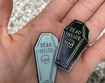 May include: Two enamel pins shaped like coffins with the text "Dead Inside" and a skull graphic. One pin is black and the other is light blue.