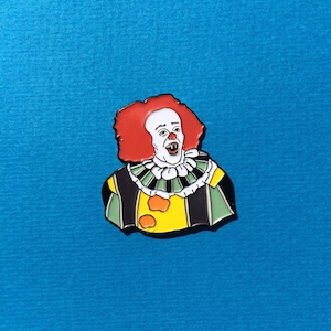 May include: A red and white enamel pin featuring a clown with a wide, toothy grin. The clown is wearing a green and black costume with a yellow collar and a red bow tie.