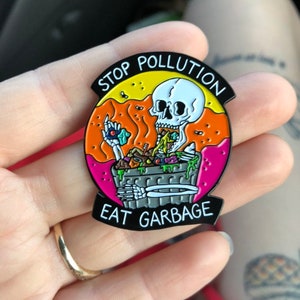 Pode incluir: Um broche de esmalte preto com um esqueleto comendo lixo de um caixote do lixo. O broche diz "Stop Pollution Eat Garbage" num c&iacute;rculo preto e branco.