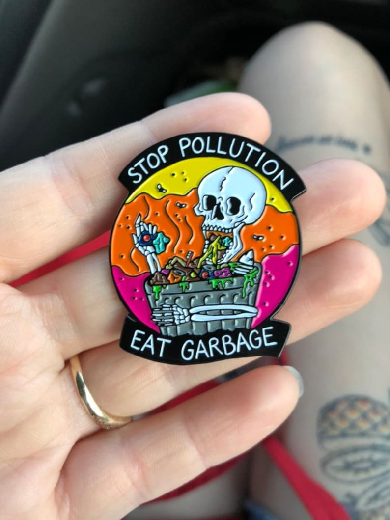 Pode incluir: Um broche de esmalte preto com um esqueleto comendo lixo de um caixote do lixo. O broche diz "Stop Pollution Eat Garbage" num c&iacute;rculo preto e branco.