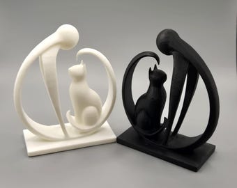 May include: Two cat sculptures, one white and one black. Each features a stylized cat figure within a curved, abstract frame. The sculptures sit on rectangular bases, creating a modern, minimalist aesthetic. The white sculpture is on the left, and the black sculpture is on the right.