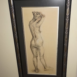 May include: A framed charcoal drawing of a nude figure. The artwork shows a person from the back, with arms raised, hair up, and legs slightly apart. The frame is dark with decorative detailing.
