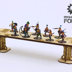 May include: A wooden display stand with eight circular slots for miniature figures. The stand is painted in a natural wood colour and has two supports on either end. The stand is displaying eight painted miniature figures of women in colourful clothing and armour. The figures are holding weapons and are posed in fighting stances. The stand is labelled "PROMETHEUM FORGE" in black text.