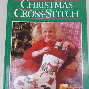 May include: A book titled "Christmas Cross-Stitch" by Better Homes and Gardens features a girl holding a red and white stocking with a cross-stitch design of Santa Claus and toys. The stocking is filled with gifts and the girl is sitting on a red and white plaid chair.