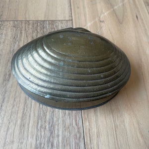 May include: A brass clam shell trinket box with a ridged design. The box is closed and sitting on a wooden surface.