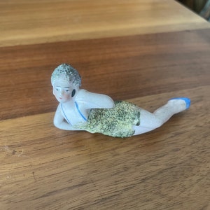 Könnte beinhalten: Eine Porzellanfigur einer Frau im Badeanzug, die auf der Seite liegt und den Arm auf dem Kopf ruht. Sie hat kurze, dunkle Haare und trägt einen blau-grünen Badeanzug.