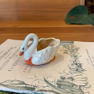 Pode incluir: Uma pequena figura de cisne de cerâmica branca com bico e pés laranja. O cisne tem a forma de um prato e tem um interior castanho. O cisne está sentado num livro com um desenho floral.