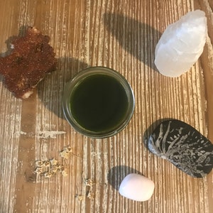 May include: A wooden table with a variety of objects on it, including a small glass jar filled with a dark green liquid, a white crystal, a pink crystal, a small brown rock, and a small pile of dried seeds.