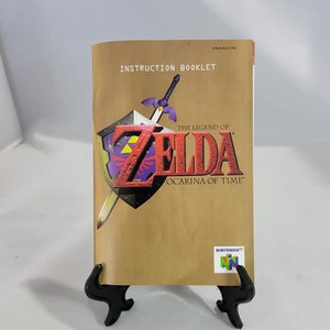May include: A brown instruction booklet for the Nintendo 64 game "The Legend of Zelda: Ocarina of Time". The cover features the game's logo with a sword and shield.