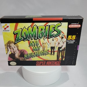 May include: A Super Nintendo game cartridge box for Zombies Ate My Neighbors. The box is black with a red and yellow border. The front of the box features a cartoon image of zombies and a woman screaming. The text on the box reads "Zombies Ate My Neighbors" and "55 Un-Deadly Levels!"
