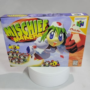 May include: A Nintendo 64 game cartridge case for the game "Mischief Makers" featuring a cartoon character in a white and red outfit with a green hat and a red glove. The character is flying in the air with a red and white jetpack. The background is a blue sky with white clouds and a group of cartoon characters in the foreground.