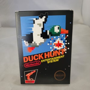 May include: A black Nintendo Entertainment System game cartridge box featuring a pixelated image of a duck flying away from a hunter. The box is labelled "Duck Hunt" and includes the Nintendo logo. The game cartridge is included.