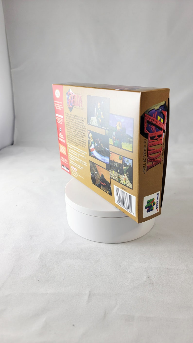 May include: A gold Nintendo 64 game cartridge case for The Legend of Zelda: Ocarina of Time. The case features a picture of Link on the front and a description of the game on the back.