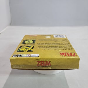 May include: The Legend of Zelda: Link's Awakening DX Game Boy Color game cartridge box. The box is gold with a picture of Link on the front. The text "The Legend of Zelda: Link's Awakening DX" is printed on the front of the box.