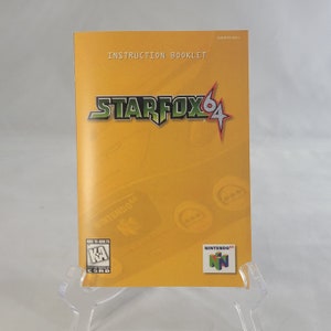 May include: A yellow instruction booklet for the video game Star Fox 64. The cover features the game's logo in green and red, with a red star. The booklet also includes the Nintendo 64 logo and a "Kids to Adults" rating from the ESRB.
