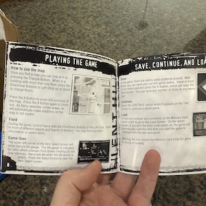 May include: Open book pages with game instructions, including "PLAYING THE GAME" and "SAVE, CONTINUE, AND LOAD" sections. The text describes map usage, saving, and loading game progress. The pages are held open by a hand.