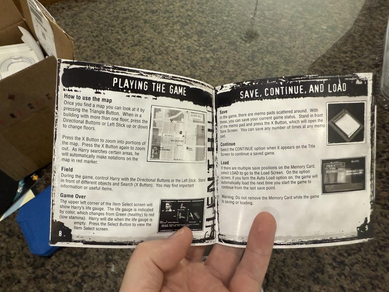 May include: Open book pages with game instructions, including "PLAYING THE GAME" and "SAVE, CONTINUE, AND LOAD" sections. The text describes map usage, saving, and loading game progress. The pages are held open by a hand.