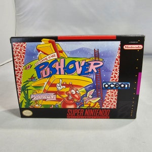 May include: A Super Nintendo Entertainment System game cartridge box for the game PushOver featuring Giant. The box is black with a red, yellow, and blue design. The game features a cartoon ant and a rabbit.