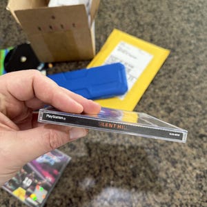 May include: A PlayStation game case for "Silent Hill" is held up, showing the game title in white text on a black spine. The case is clear plastic, and the PlayStation logo is visible. Other items are in the background.