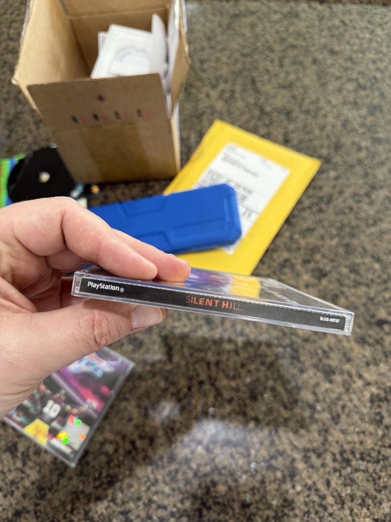 May include: A PlayStation game case for "Silent Hill" is held up, showing the game title in white text on a black spine. The case is clear plastic, and the PlayStation logo is visible. Other items are in the background.