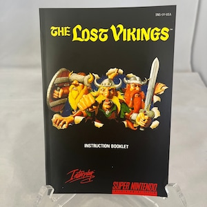 May include: The Lost Vikings Super Nintendo Entertainment System instruction booklet cover features three cartoon Viking warriors with axes and swords, breaking through a black background. The title "The Lost Vikings" is in yellow text.
