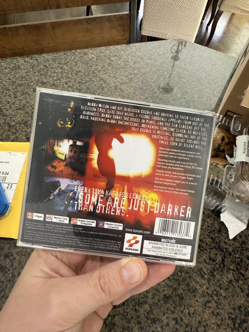 May include: A video game case with the title "Silent Hill" is held in a hand. The cover art features a silhouetted figure and the text "Every town has its secrets, some are just darker than others."