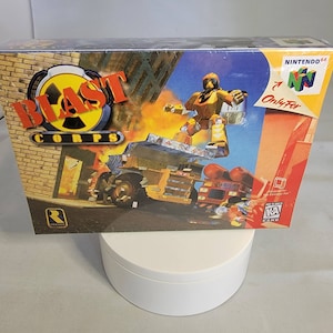 Peut inclure: Boîte de jeu vidéo Blast Corps pour Nintendo 64. La boîte présente un personnage de robot jaune debout sur un bâtiment avec un camion au premier plan. Le titre du jeu est en lettres rouges et noires. La boîte est en bon état avec quelques signes d'usure.