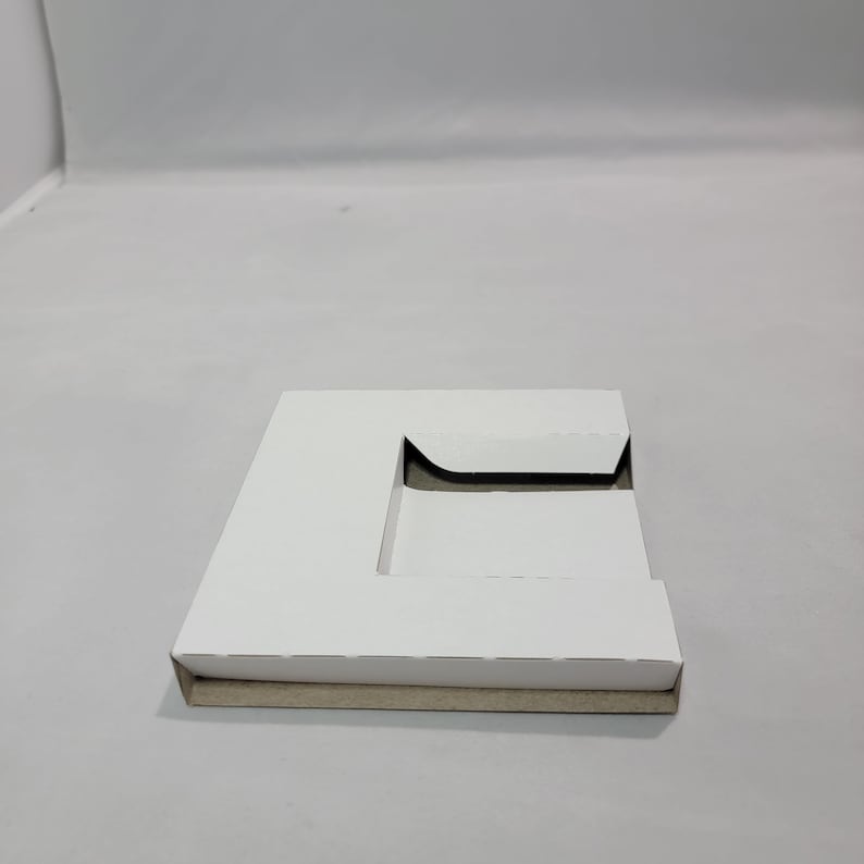 May include: White cardboard box with a cut-out shape in the center. The box is folded and has a brown cardboard bottom.
