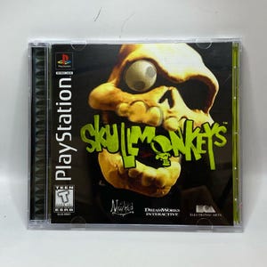 Peut inclure: Boîtier de jeu PlayStation pour "Skullmonkeys" avec un crâne grotesque avec un grand œil. Le titre du jeu est en vert vif, avec le logo PlayStation en haut à gauche. Le boîtier est en plastique transparent.