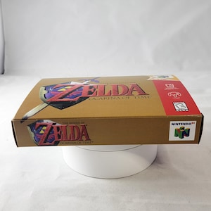 May include: A gold Nintendo 64 game cartridge case for The Legend of Zelda: Ocarina of Time. The case features a red and gold design with the game's logo and title.