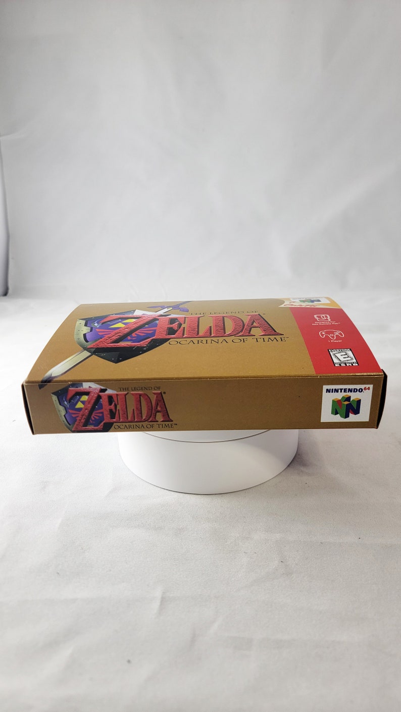 May include: A gold Nintendo 64 game cartridge case for The Legend of Zelda: Ocarina of Time. The case features a red and gold design with the game's logo and title.