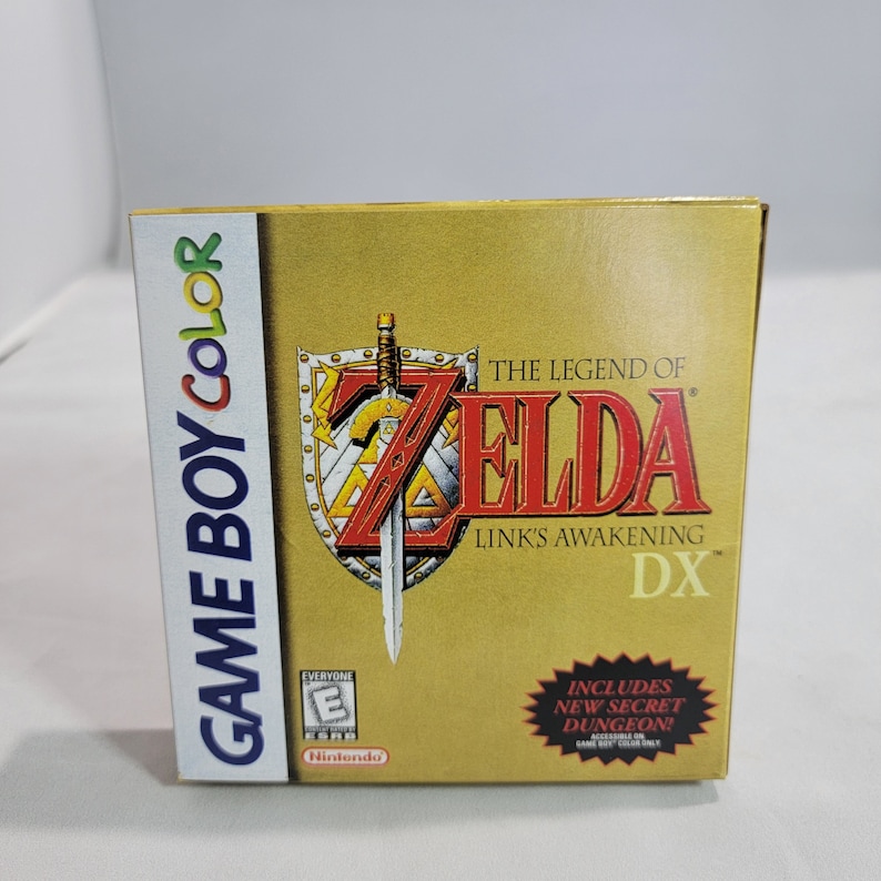 May include: A gold Game Boy Color game cartridge for The Legend of Zelda: Link's Awakening DX. The cartridge features a red and gold shield with a sword and the game title.