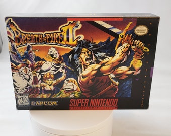 Puede incluir: Una caja negra de cartucho de juego Super Nintendo con una imagen colorida de un hombre con el pelo largo y una espada, junto con otros personajes. El juego se llama "Breath of Fire II" y está hecho por Capcom.