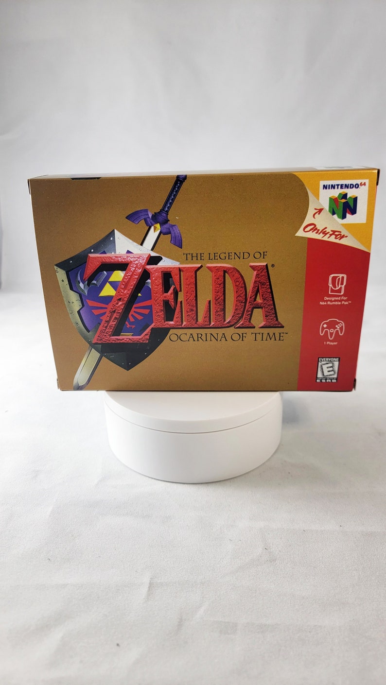 May include: A gold Nintendo 64 game cartridge case for The Legend of Zelda: Ocarina of Time. The case features a large image of Link and the Triforce, along with the game's title and logo. The case is designed for the Nintendo 64 console and is rated E for Everyone by the ESRB.