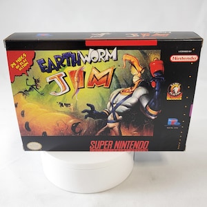 May include: A black Super Nintendo game cartridge box with a colorful image of a cartoon worm character wearing a blue and white suit and holding a red weapon. The box has the title "Earthworm Jim" and the text "24 megs of play action".