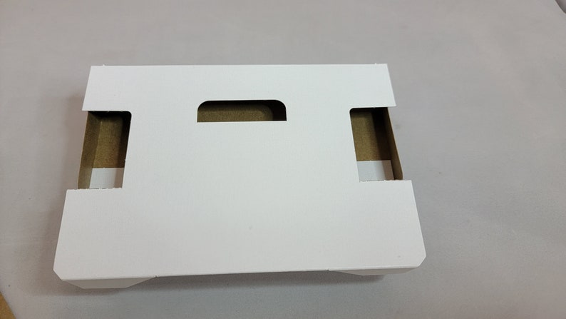 May include: White cardboard box with two cut-out handles. The box is designed to hold and protect items during shipping.